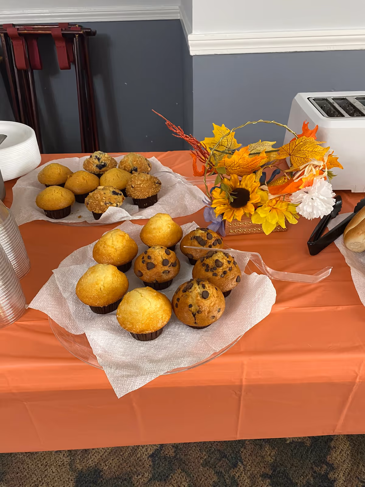 Table covered with an orange tablecloth displaying plates of assorted muffins including plain, blueberry, and chocolate chip varieties, a decorative autumn-themed floral arrangement, a white toaster, plastic cups, and plates stacked on the side.