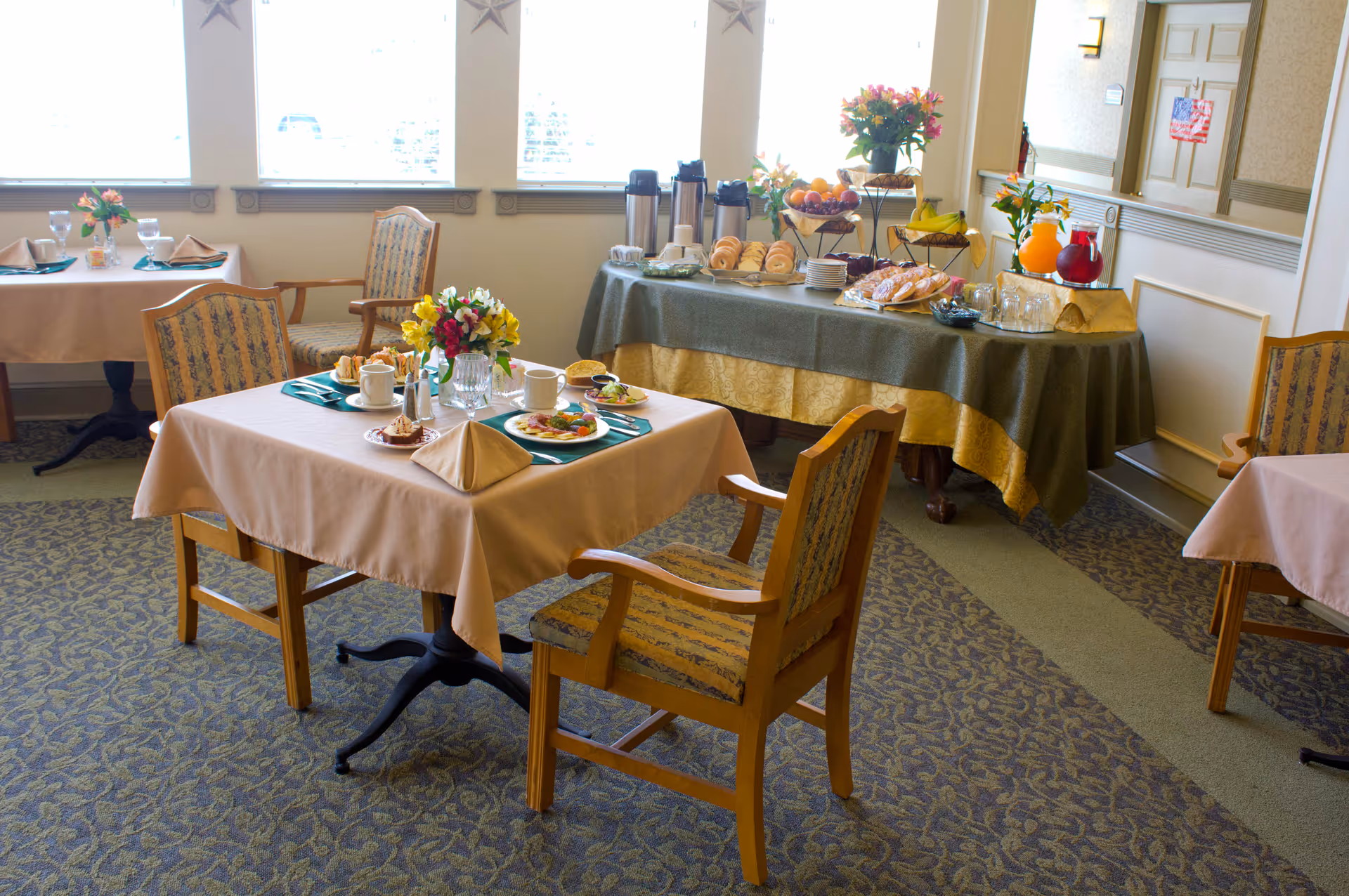 A dining room with tables covered in beige tablecloths, set with plates of food, cups, and napkins. There are wooden chairs with patterned cushions around the tables. In the background, a buffet table is set with coffee dispensers, plates, pastries, fruit, and pitchers of juice. The room has large windows letting in natural light.