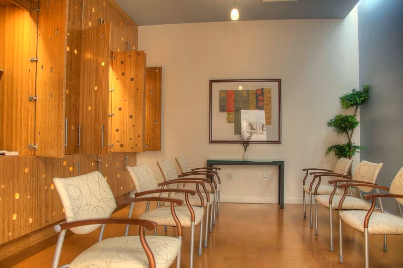 A waiting room with two rows of beige cushioned chairs with wooden armrests facing each other. On the left wall, there are wooden cabinets with circular cut-out designs. At the far end of the room, a small table holds a framed picture and a decorative item, with a framed abstract artwork hanging above it. A potted plant is placed in the corner on the right side. The room has a warm, inviting atmosphere with soft lighting.
