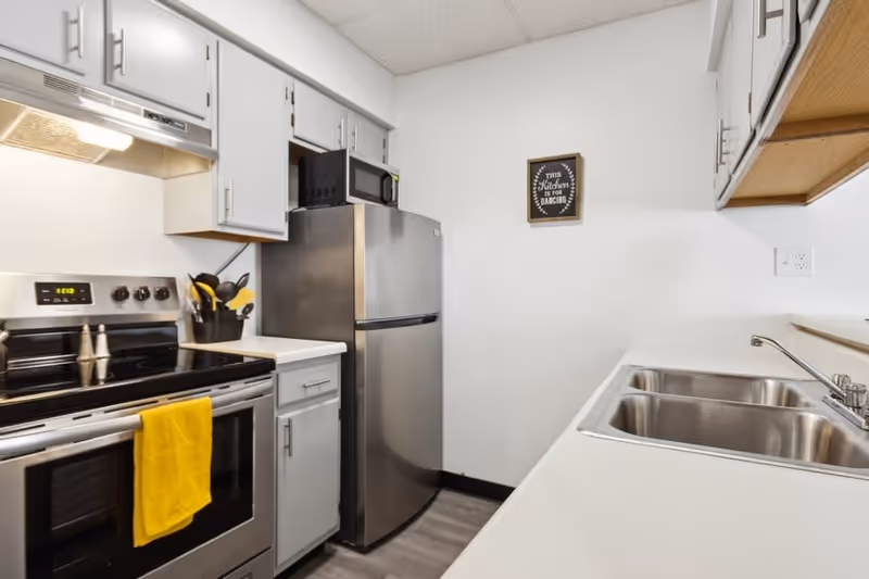 A modern kitchen with white cabinets, a stainless steel refrigerator, stove, and microwave. There is a double sink on the right side and a yellow towel hanging on the oven handle. A small framed sign on the wall reads 'This kitchen is for dancing.'