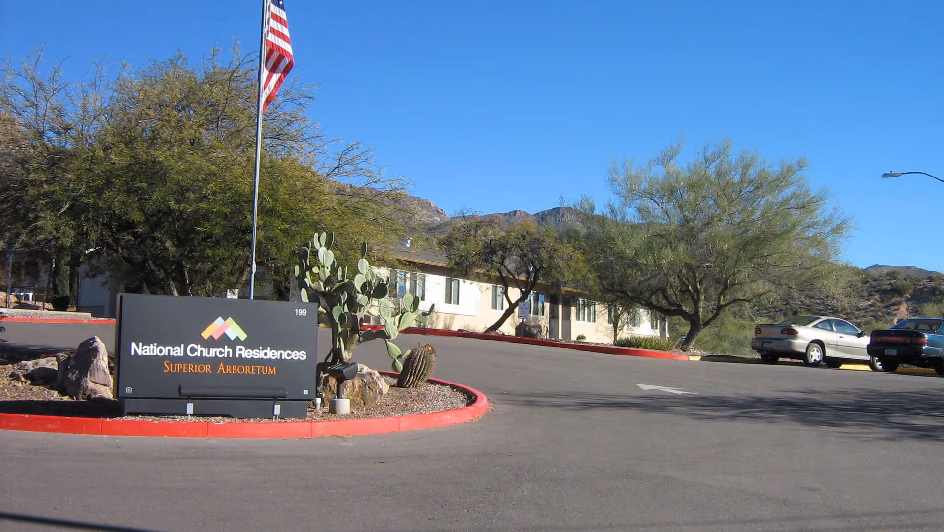 Exterior view of the National Church Residences Superior Arboretum facility with a sign in the foreground, an American flag on a pole, desert landscaping including cacti and trees, a paved driveway, parked cars, and mountains in the background under a clear blue sky.