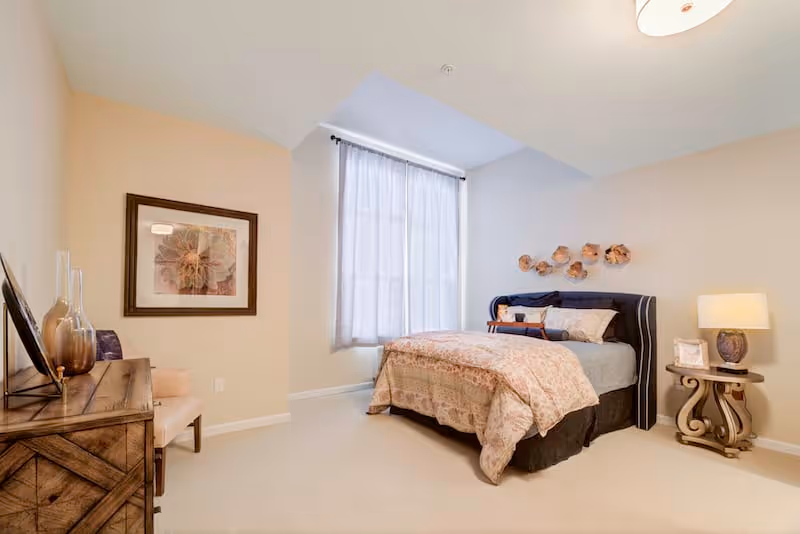 A bright bedroom with a large window covered by sheer curtains, a bed with patterned bedding and decorative pillows, a nightstand with a lamp and framed photo, a wooden dresser with decorative vases, a chair, and a framed floral artwork on the wall.