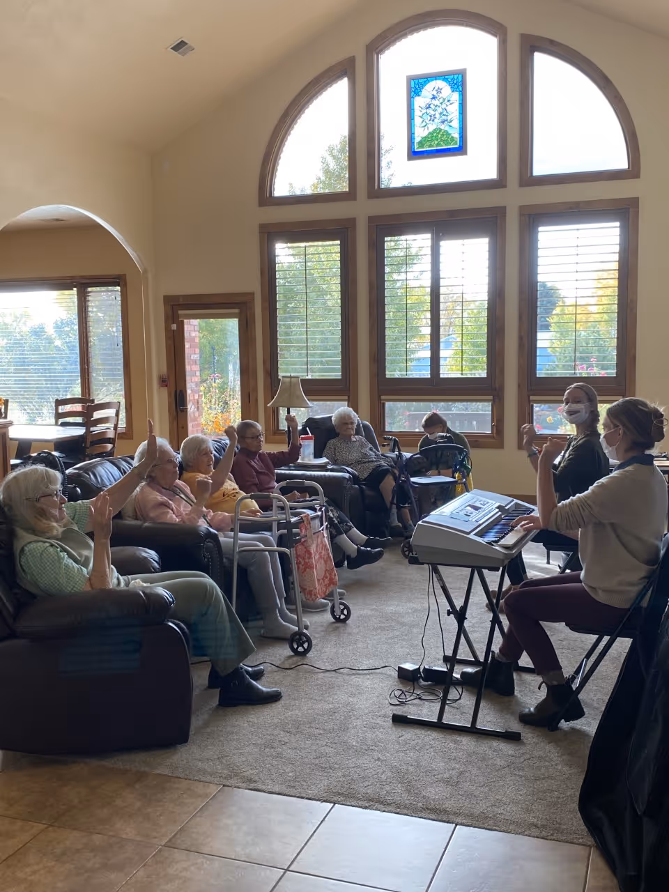 A group of seniors seated in a sunny common room with large arched windows participating in a music activity while a staff member plays a keyboard.