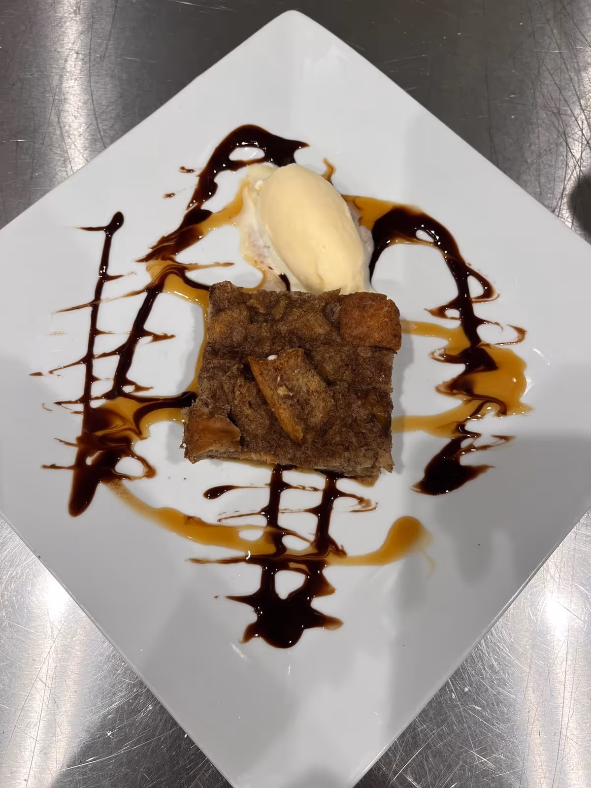 A square piece of dessert, possibly bread pudding, served on a white square plate with a scoop of vanilla ice cream and drizzles of caramel and chocolate sauce on the plate.