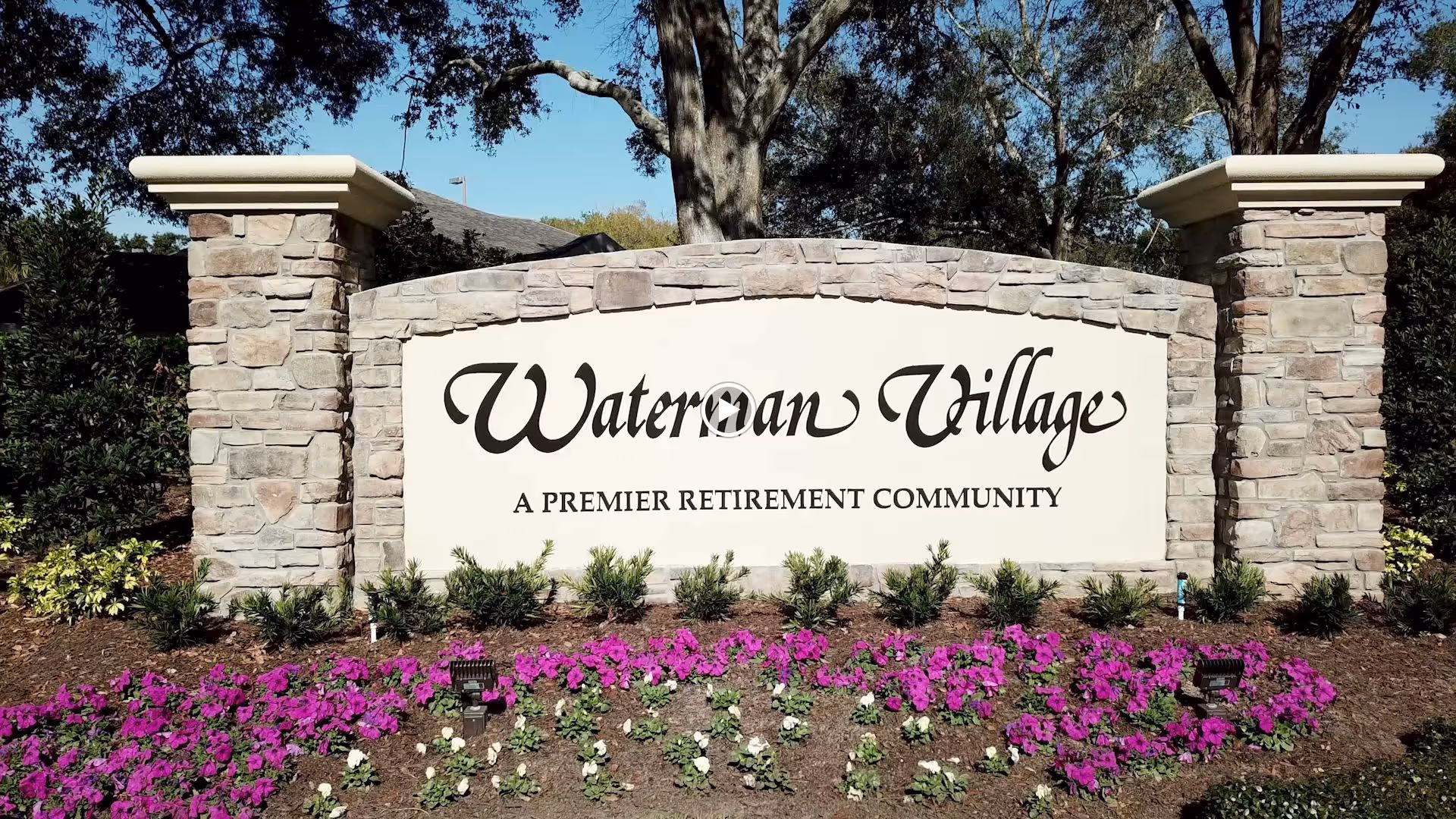 Stone entrance sign for Waterman Village, a premier retirement community, surrounded by landscaping with purple and white flowers and greenery under a clear blue sky.