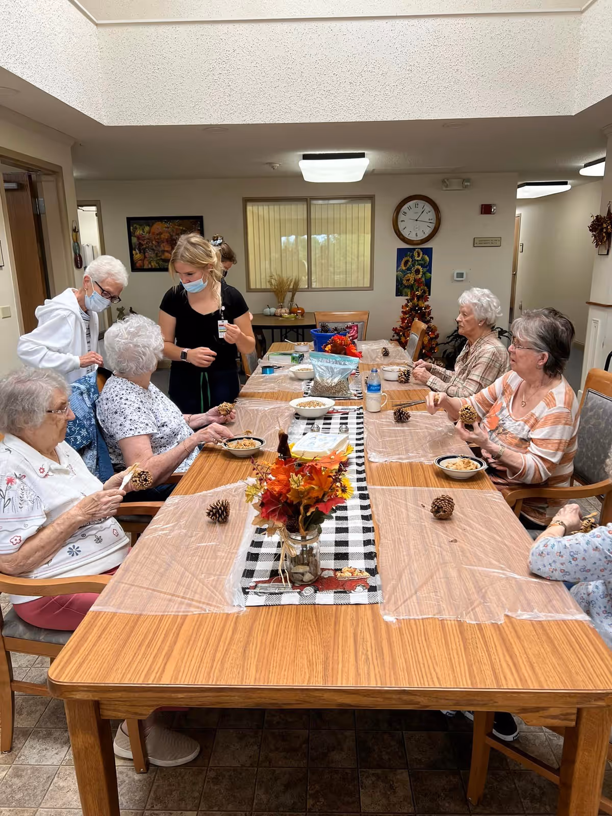 Several elderly residents and a masked staff member sit around a long table doing a pinecone craft in a communal dining/activity room.