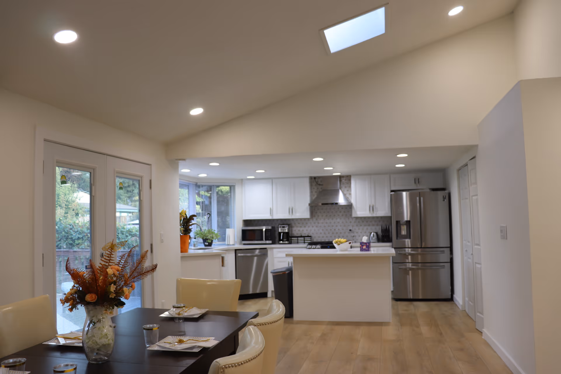 A bright, modern kitchen and dining area with white cabinets, stainless steel appliances including a refrigerator and dishwasher, a kitchen island with a bowl of fruit, and a dining table with cream-colored chairs and a floral centerpiece. There are large windows and glass doors letting in natural light, and recessed ceiling lights illuminate the space.