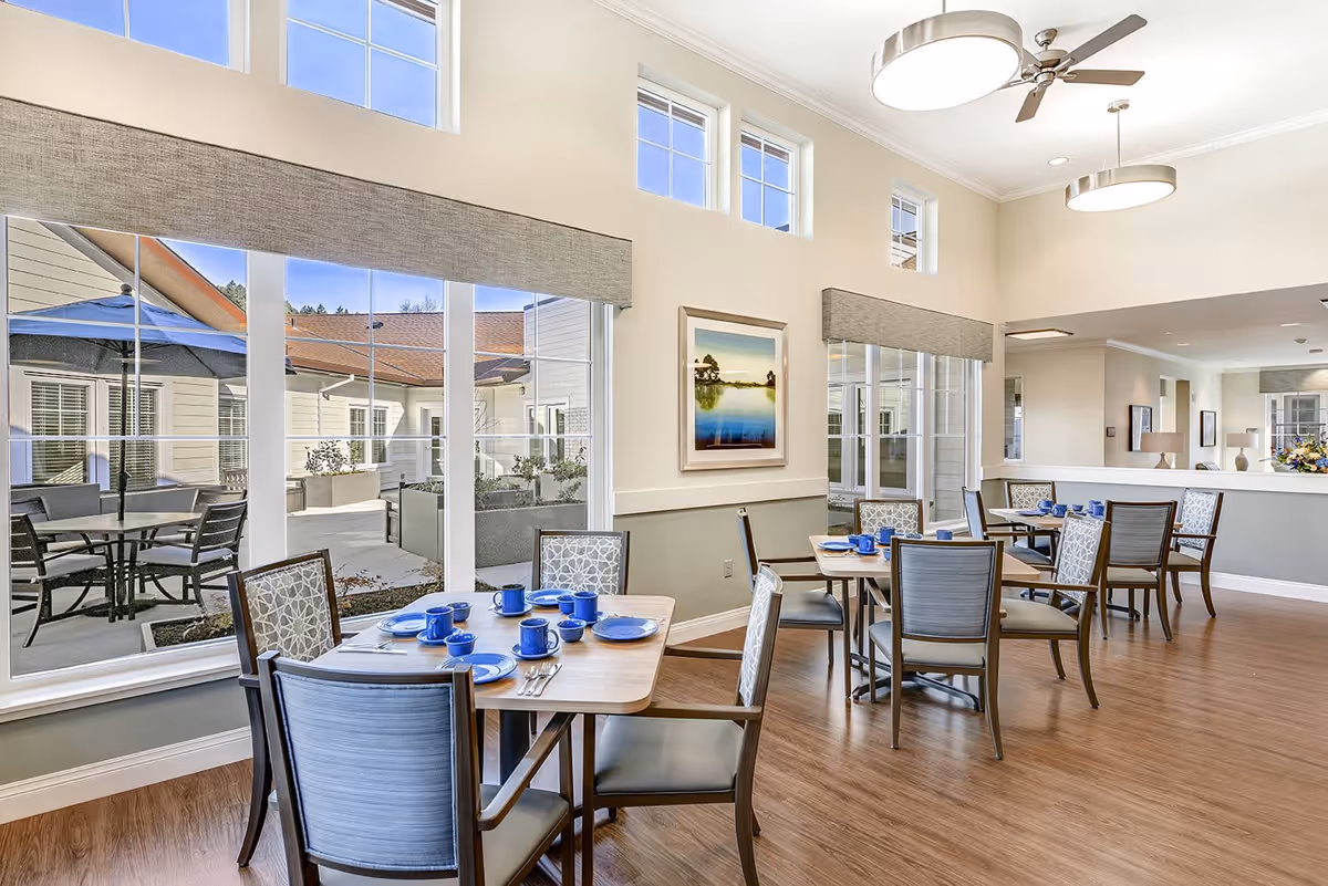 A bright dining room in a senior living facility with several wooden tables set with blue plates, cups, and silverware. The room has large windows with views of an outdoor patio area with tables and umbrellas. The walls are light-colored, and there are modern ceiling lights and a ceiling fan. A framed landscape painting hangs on the wall.