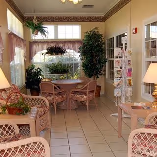 Sunlit common room with wicker seating and a small dining table surrounded by plants and a greeting card rack.