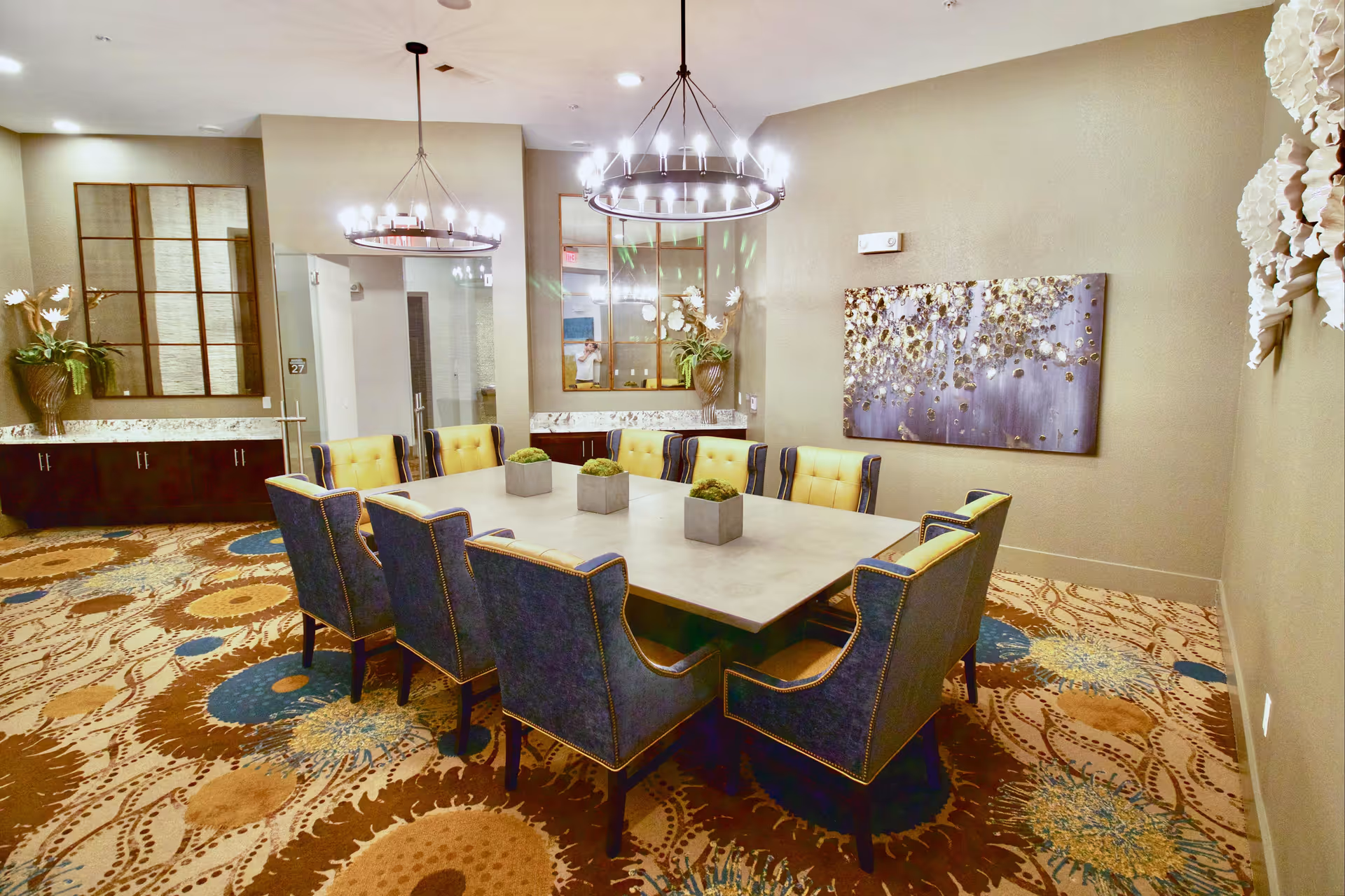 A well-lit dining room with a large rectangular table surrounded by twelve upholstered chairs in blue and yellow. The room features patterned carpet, two circular chandeliers hanging from the ceiling, decorative wall art, and large mirrors on the walls. There are also plants and a cabinet along one wall.