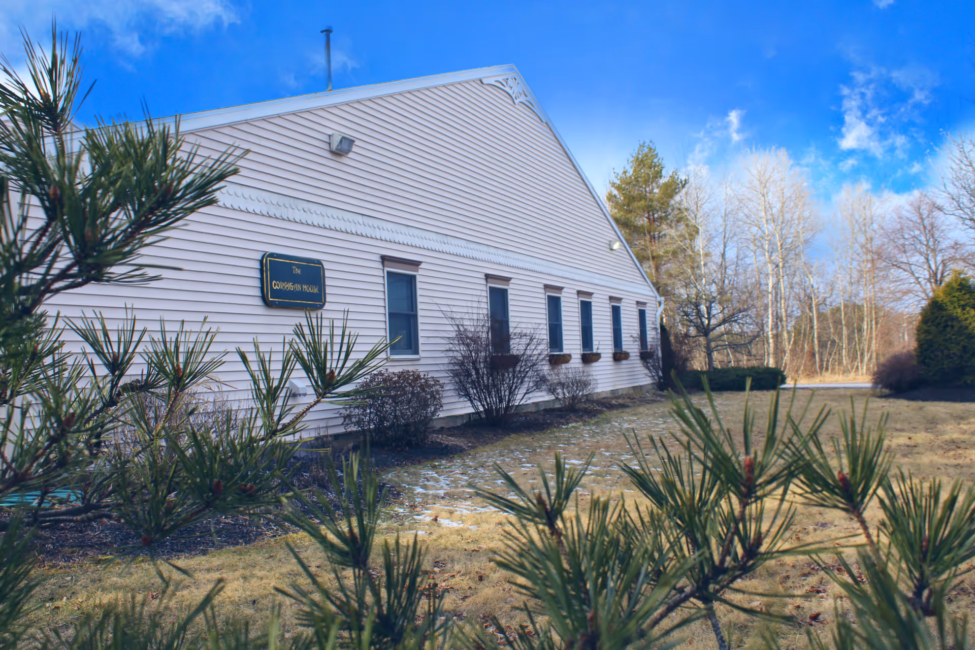 Exterior view of a white building with horizontal siding, several windows with flower boxes, and a sign that reads 'The Corrigan House'. The foreground includes green pine branches and a grassy area with some bushes and trees in the background under a blue sky with scattered clouds.