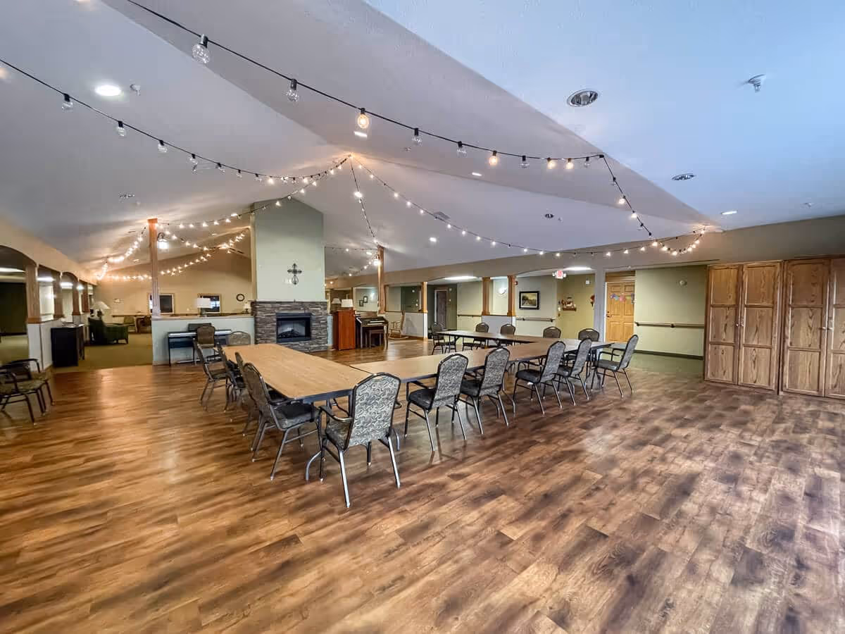 A spacious common area in an assisted living facility with wooden flooring, several tables arranged in a U-shape surrounded by chairs, a stone fireplace in the center, string lights hanging from the ceiling, and wooden cabinets along one wall.