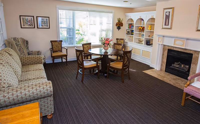 A cozy living room area with a patterned sofa and armchair on the left, a round wooden table with six chairs in the center, a fireplace on the right, and a built-in white bookshelf decorated with various items. A large window with blinds lets in natural light, and framed pictures hang on the walls.