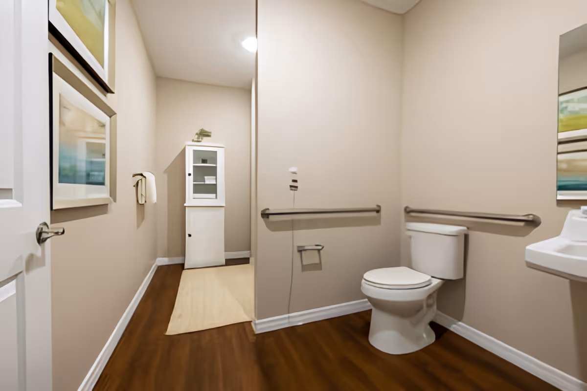 Accessible bathroom with a toilet, grab bars, sink, and a small white storage cabinet in a beige room.
