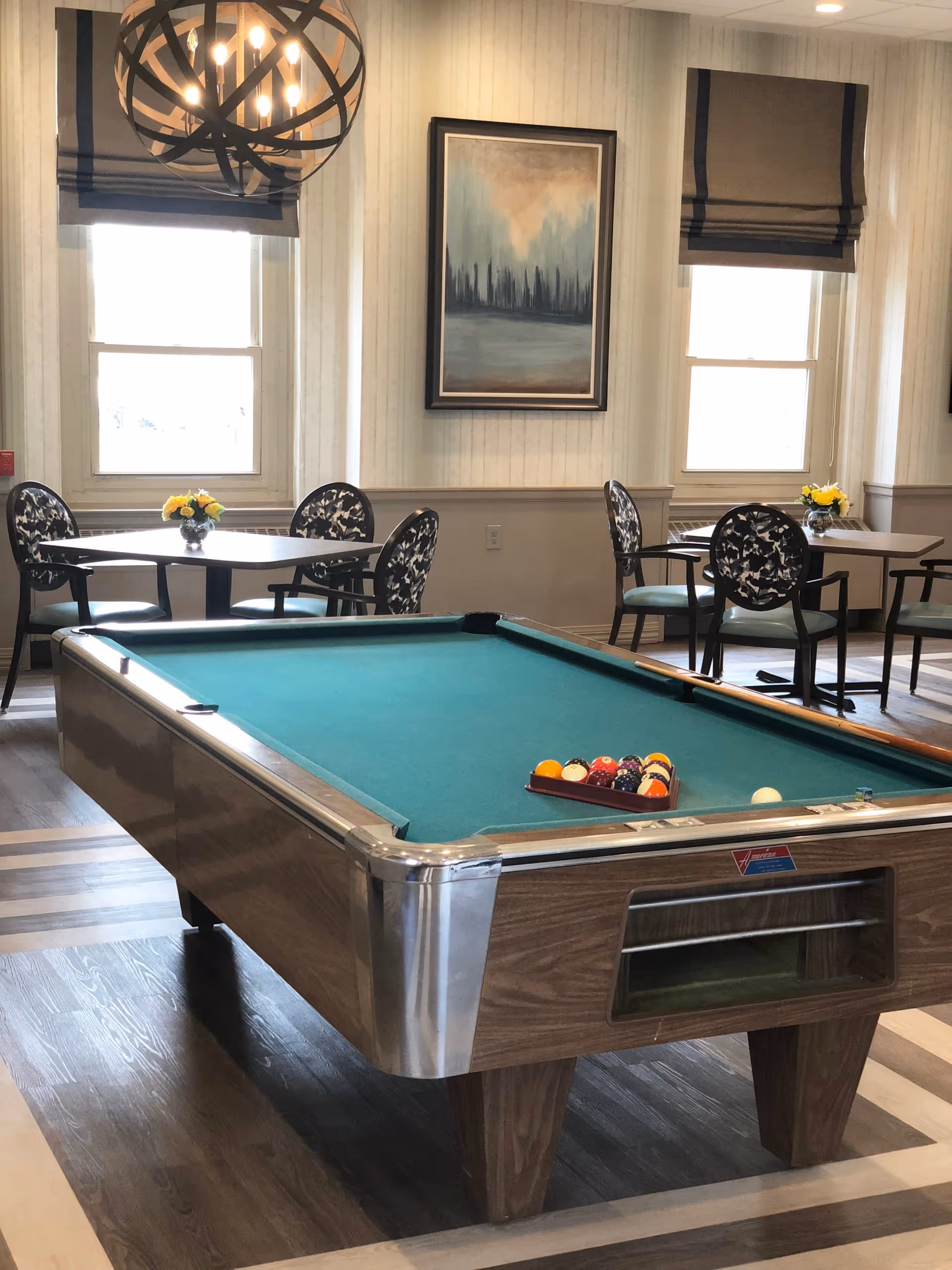 Interior room with a pool table in the foreground and two small dining tables with chairs in the background. The room has two windows with brown Roman shades, a modern spherical chandelier, and a framed abstract painting on the wall. Each dining table has a small vase with yellow flowers.