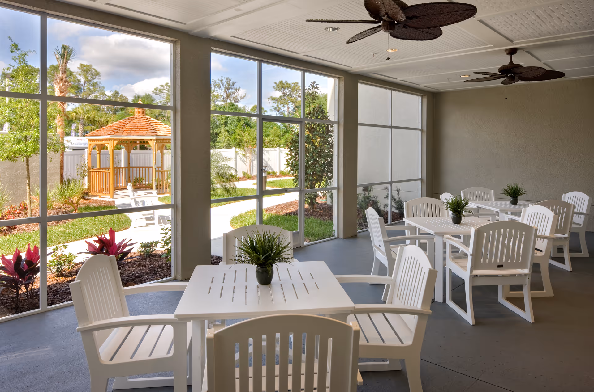 Covered outdoor seating area with white tables and chairs overlooking a garden and a wooden gazebo.