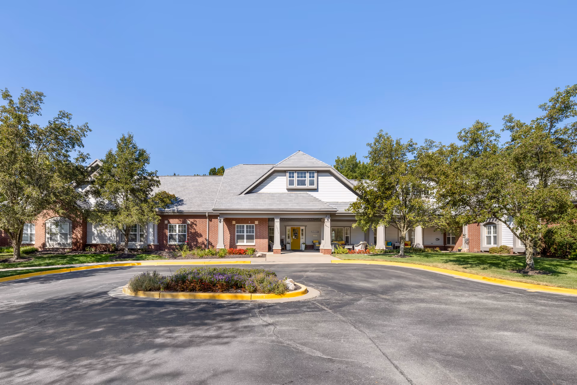 Front exterior view of Brookdale Leawood State Line facility with a circular driveway, landscaped garden in the center, trees on both sides, and a clear blue sky.