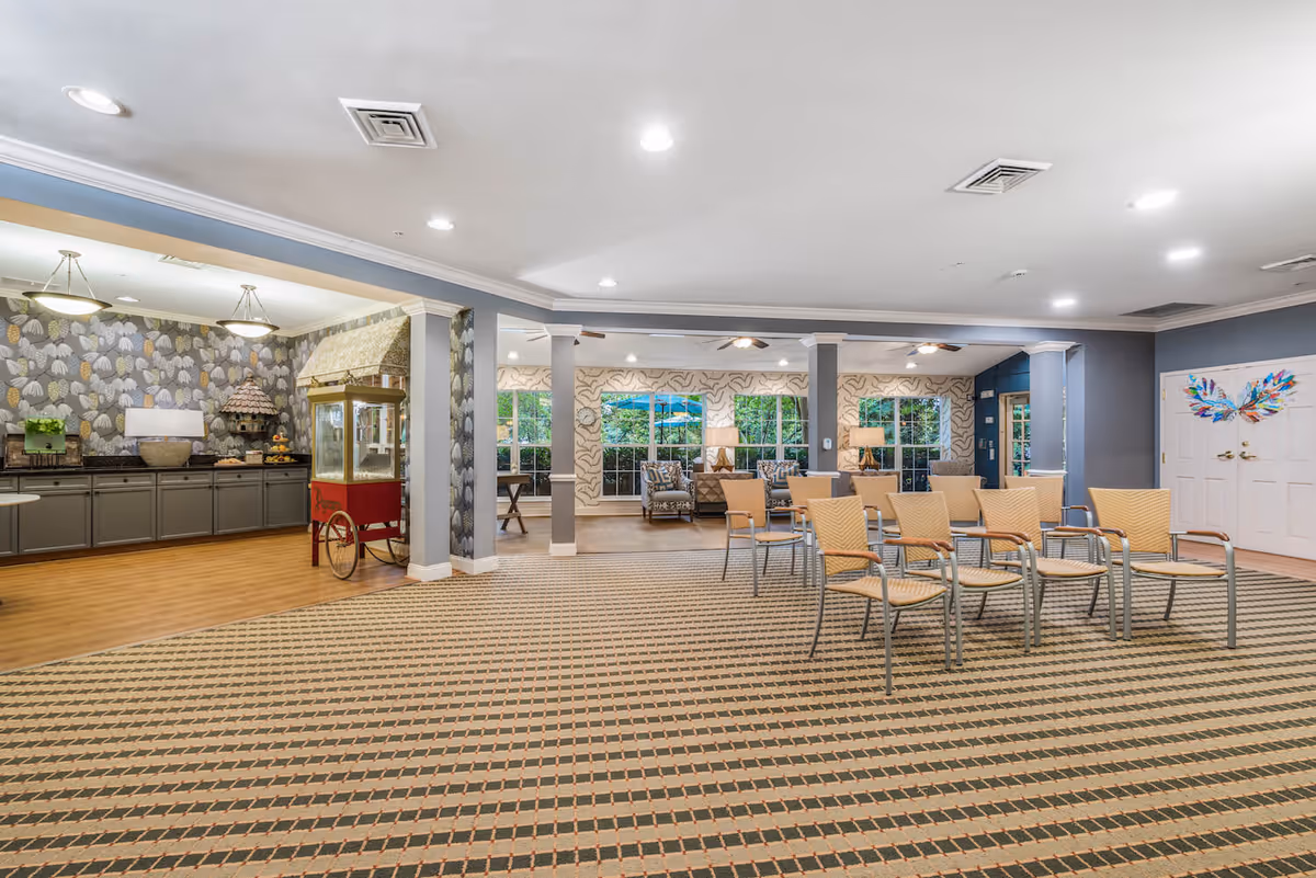A spacious common area in a senior living facility with a patterned carpet and several beige chairs arranged in rows. The room features a popcorn machine on the left side near a counter with cabinets and decorative wallpaper. In the background, there are large windows with views of greenery outside, and a seating area with armchairs and lamps. The walls are painted in soft blue and white tones, and the ceiling has recessed lighting.