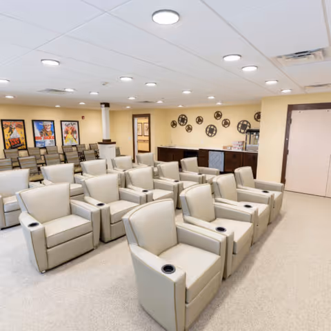 A senior living facility theater room with multiple rows of beige recliner chairs equipped with cup holders, a few rows of standard chairs at the back, movie posters on the wall, and decorative film reels mounted on a yellow wall.