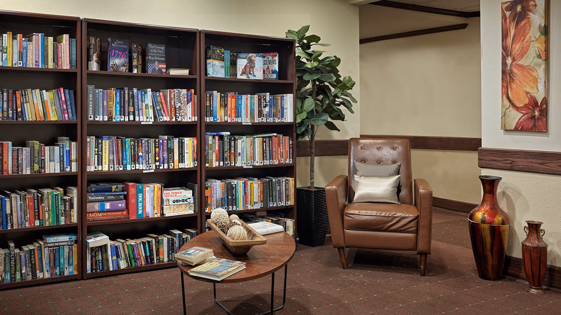 A cozy reading nook in a senior living facility featuring three dark wood bookshelves filled with books, a round wooden coffee table with decorative items and books on it, a brown leather armchair with a beige pillow, a tall green potted plant, and two large decorative vases next to a wall with floral artwork.