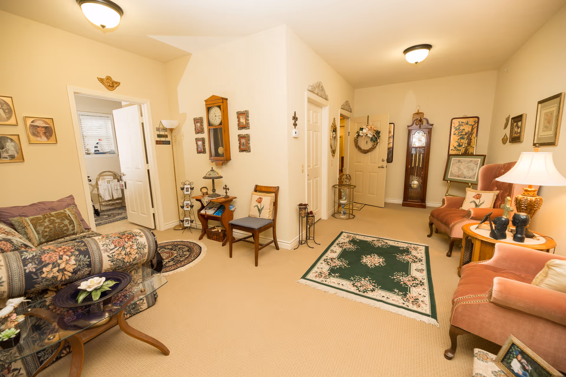 A cozy living room area with beige walls and carpet, featuring floral patterned sofa and two pink upholstered armchairs. There are decorative rugs on the floor, a wooden grandfather clock, framed pictures on the walls, and various small tables with lamps and decorative items. Two open doors lead to other rooms, one showing a glimpse of a bed and window.