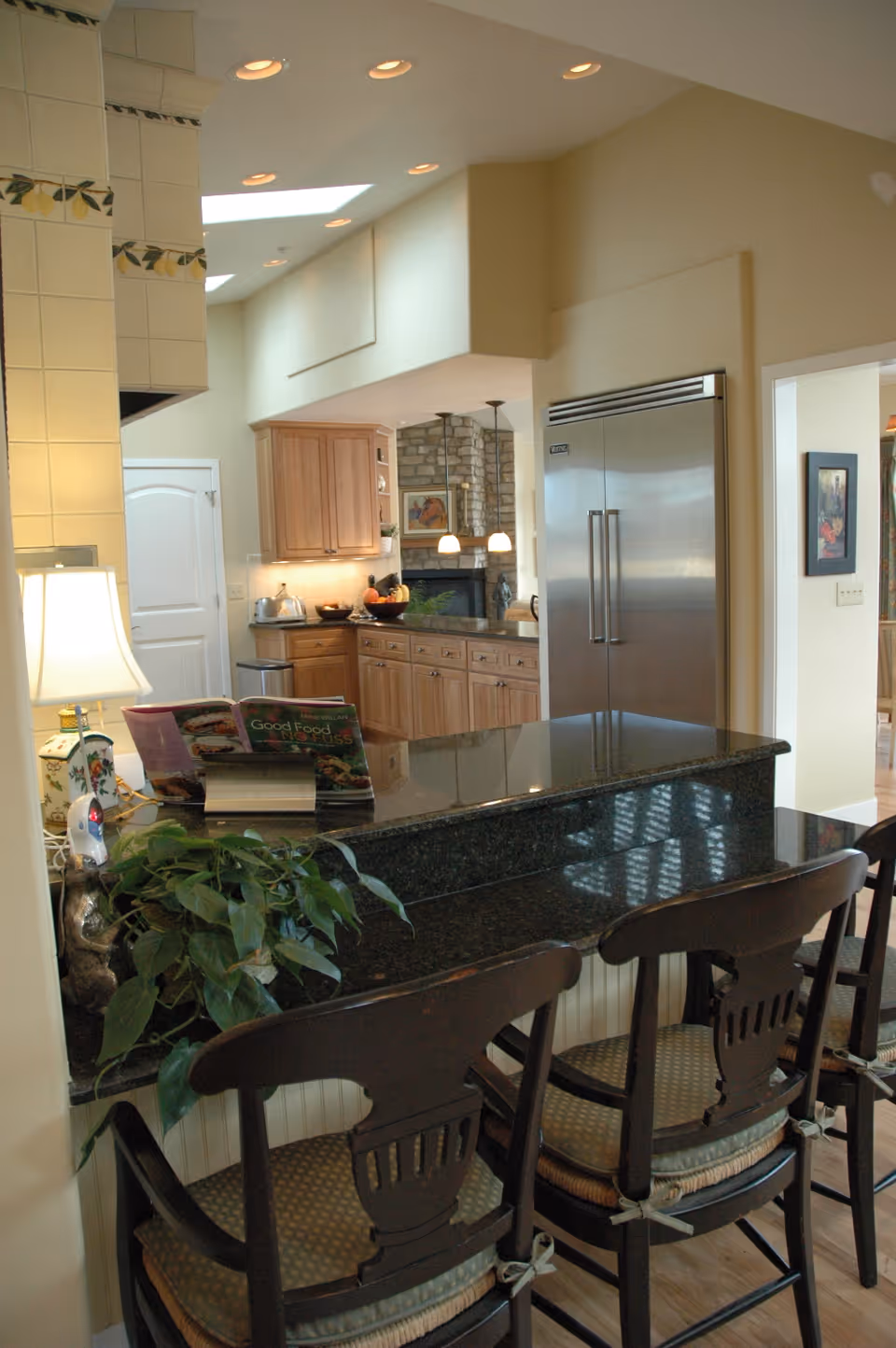Interior view of a kitchen area featuring a black granite countertop with three wooden chairs with cushions. The kitchen has wooden cabinets, a large stainless steel refrigerator, and a tiled wall with decorative accents. There is a plant and a lamp on the counter, and a cookbook is open on a stand. The background shows a stone fireplace and pendant lights in an adjoining room.