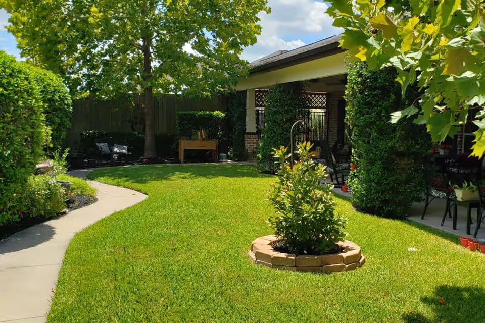 A well-maintained outdoor garden area with a green lawn, a small circular flower bed with a shrub in the center, a curved concrete pathway, and a covered patio with chairs and greenery-covered pillars. Trees and bushes surround the space under a partly cloudy sky.