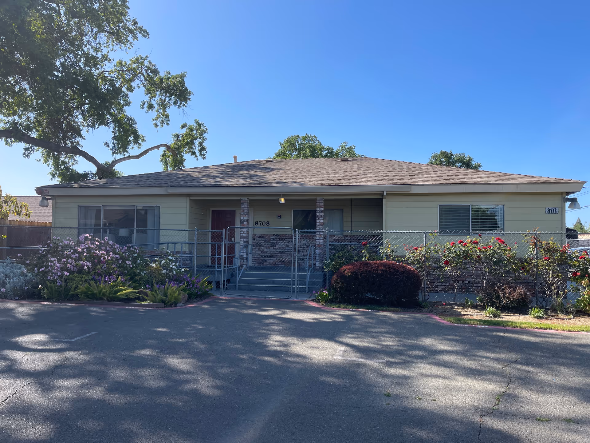Single-story yellow building front with a fenced porch, address number 8708, and landscaped flower beds.