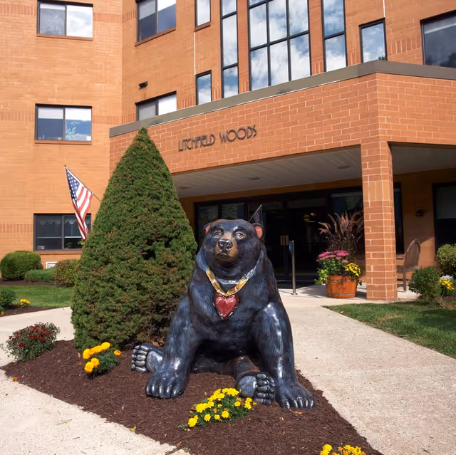 Entrance of a brick building named Litchfield Woods with a large black bear statue wearing a heart-shaped pendant in front, surrounded by flowers and a neatly trimmed conical bush. An American flag is visible to the left of the entrance.