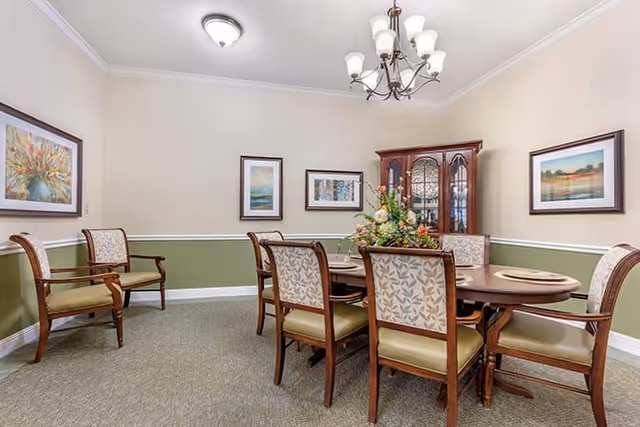 A dining room with a wooden oval table surrounded by six upholstered chairs. The table is set with placemats and a floral centerpiece. The walls are painted beige with a green wainscoting and decorated with framed landscape paintings. A wooden china cabinet is against the far wall, and a chandelier with multiple lights hangs from the ceiling.