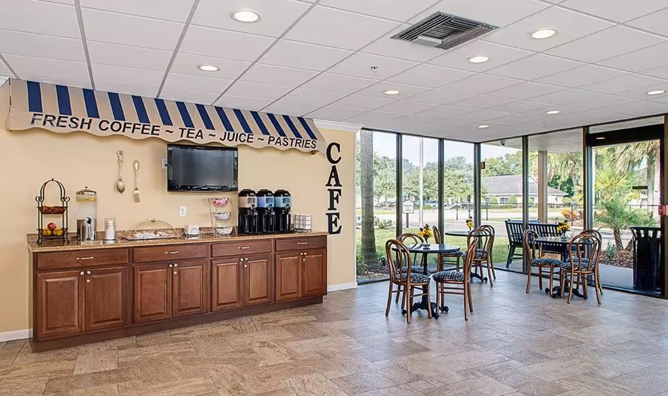 A bright cafe area with large windows showing an outdoor view. Inside, there are several small round tables with chairs, and a counter with coffee dispensers, juice, pastries, and a water dispenser. Above the counter is a striped awning with the words 'FRESH COFFEE ~ TEA ~ JUICE ~ PASTRIES' and the word 'CAFE' vertically on the wall.