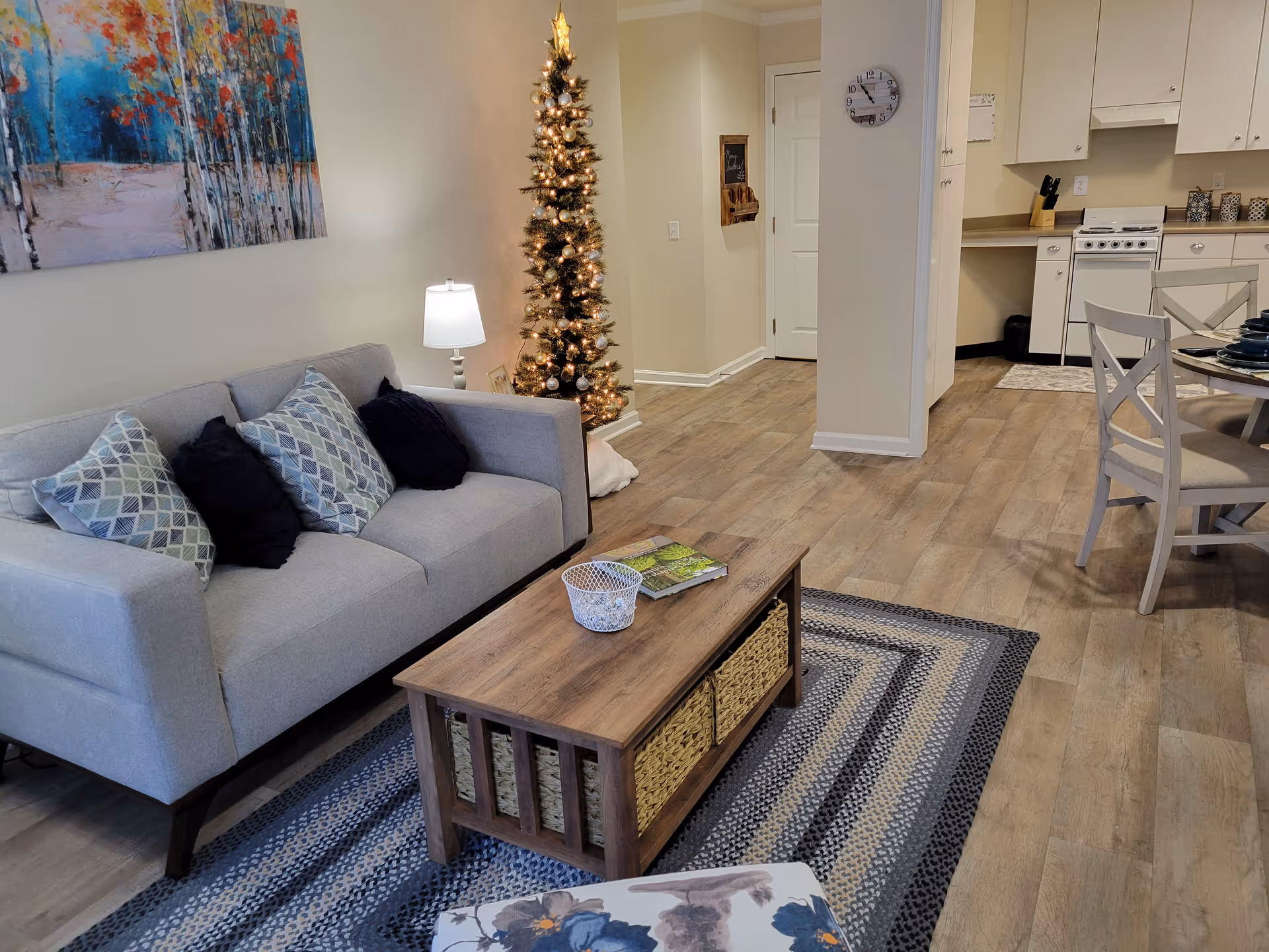 A cozy living room area with a light gray sofa adorned with patterned and solid pillows, a wooden coffee table with woven baskets underneath, and a patterned rug. A decorated Christmas tree with lights stands next to a small table lamp. In the background, there is a kitchen area with white cabinets, a stove, and a dining table with chairs. A wall clock and a small chalkboard are visible on the wall near a closed door.