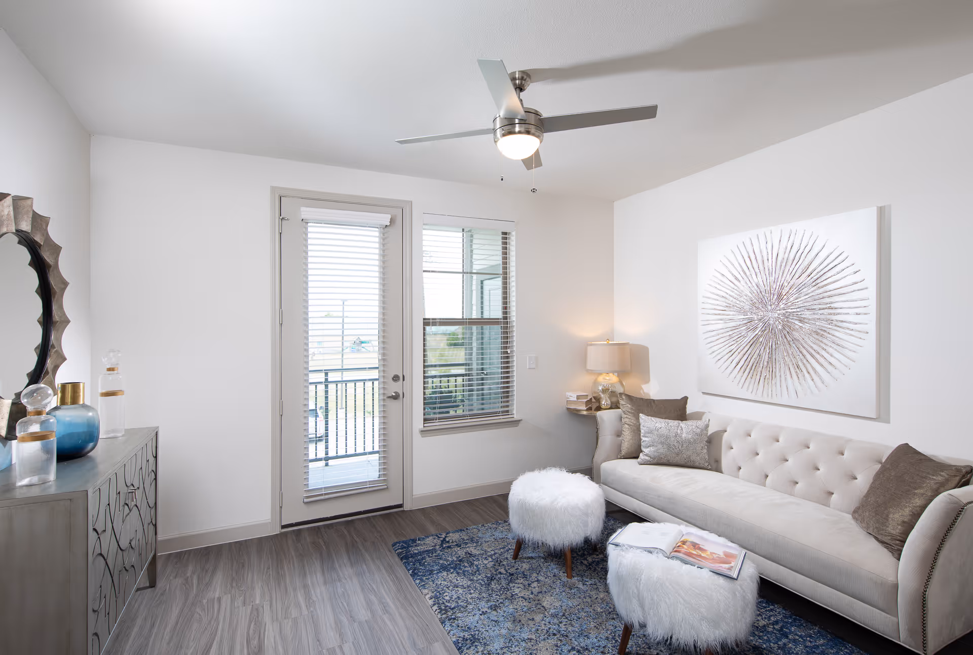 Bright modern living room with a tufted sofa, two fluffy ottomans, decorative console, artwork, and a glass door opening to a balcony.