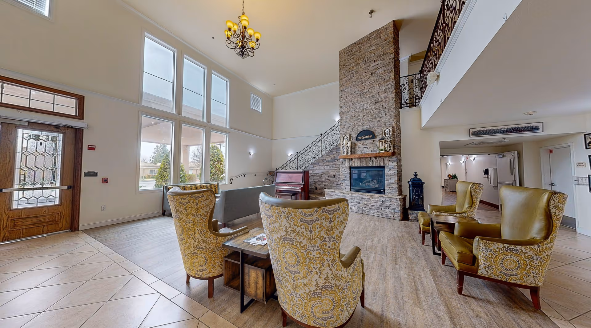 A spacious and well-lit living room area with high ceilings and large windows letting in natural light. The room features a stone fireplace with a 'welcome' sign above it, a piano, and several upholstered armchairs arranged around small tables. There is a wooden door with decorative glass on the left and a staircase with wrought iron railing leading to an upper level.