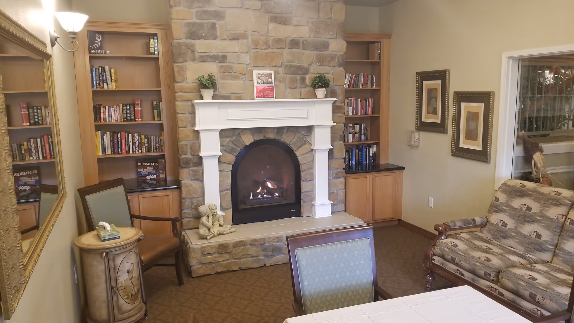 Cozy living room area with a stone fireplace in the center, flanked by built-in wooden bookshelves filled with books. A small statue sits on the hearth. There are several chairs and a patterned sofa around a table with a white tablecloth. A large mirror hangs on the left wall, and two framed pictures are on the right wall. A window is visible on the right side.