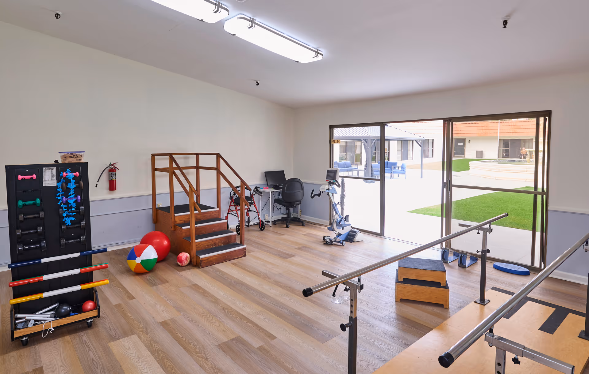 A rehabilitation room with wooden flooring featuring parallel bars for walking exercises, a small set of wooden stairs with handrails, various exercise equipment including colorful balls, dumbbells, and resistance bands. There is a desk with a chair and computer near large sliding glass doors that open to an outdoor courtyard with seating and green grass.