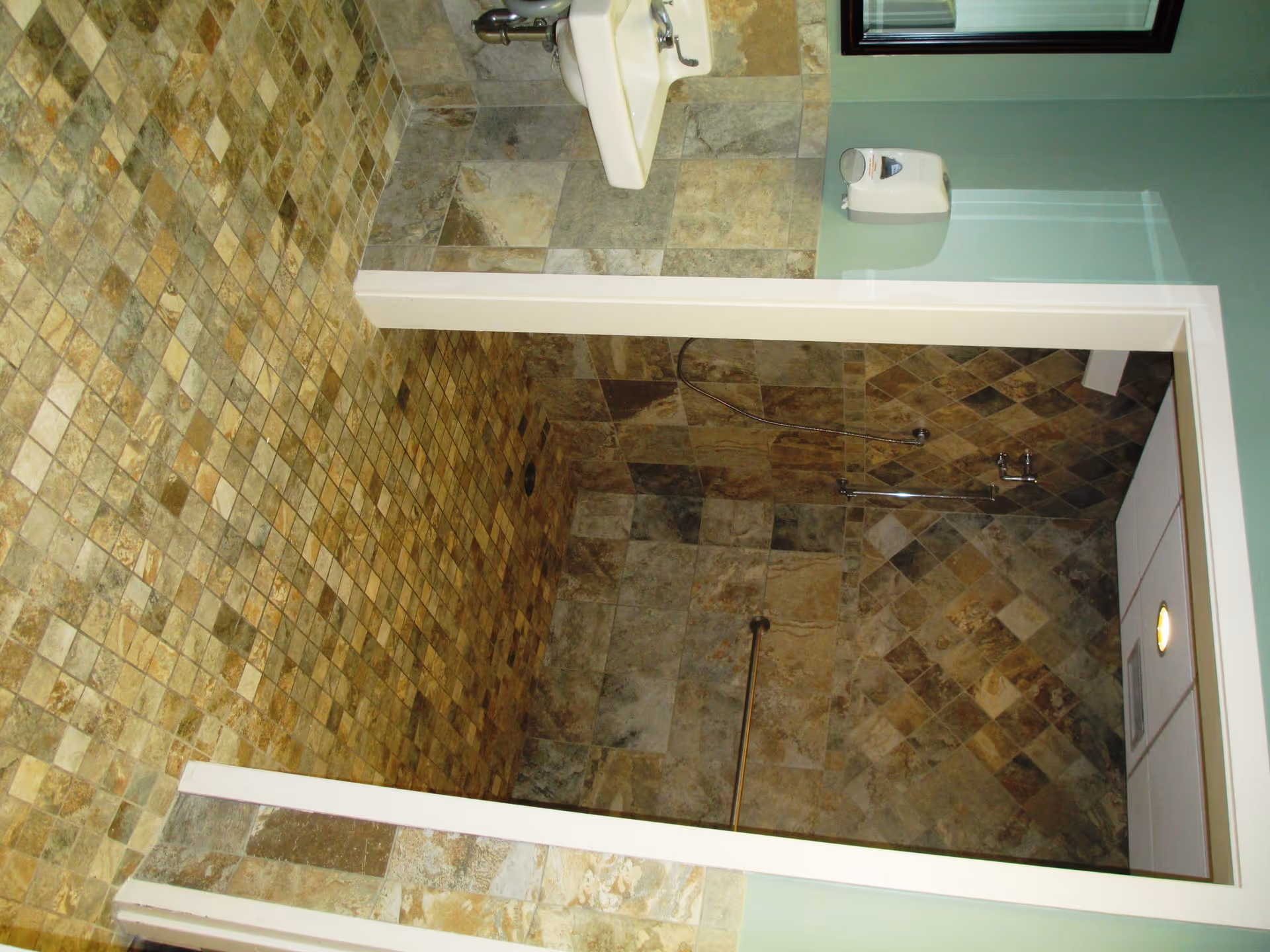 A tiled bathroom with a walk-in shower area featuring a handheld showerhead and grab bar. The floor and walls are covered with square stone tiles in various shades of brown and beige. A white sink is mounted on the wall outside the shower, and a soap dispenser is attached to the green-painted wall above the sink.