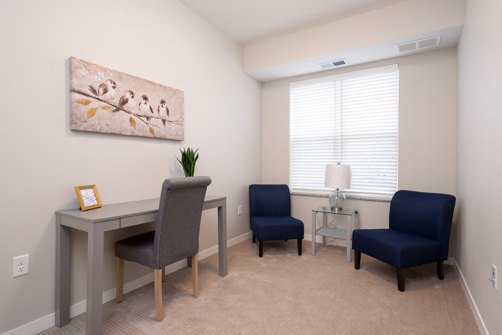 Small sitting room with a desk and chair on the left, two blue armchairs and a glass side table with a lamp by a window, and bird artwork on the wall.