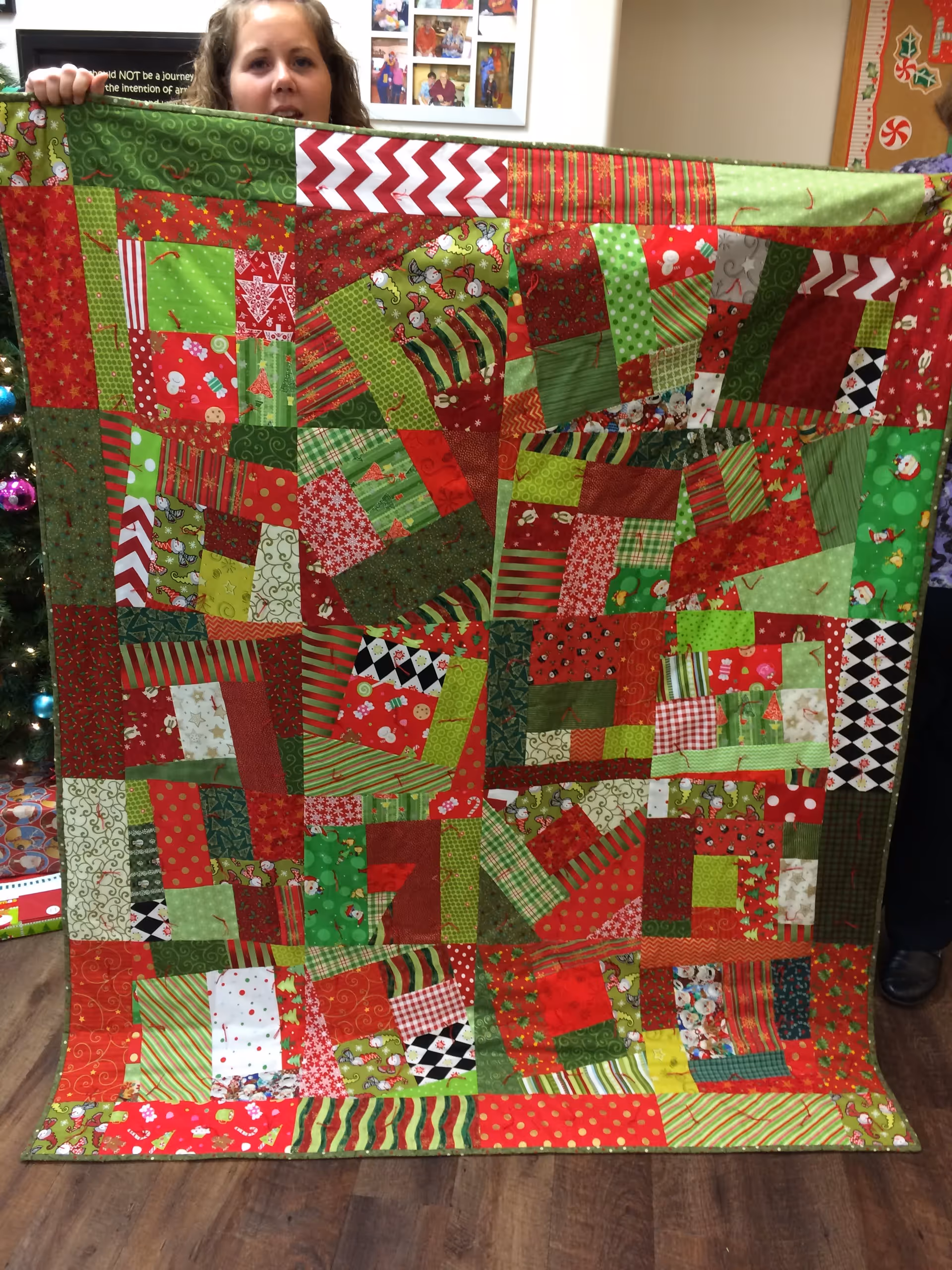 A person holding up a colorful patchwork quilt with various red, green, and patterned fabric squares. The quilt has a festive, holiday theme with Christmas-related designs. In the background, there is a decorated Christmas tree and framed photos on the wall.