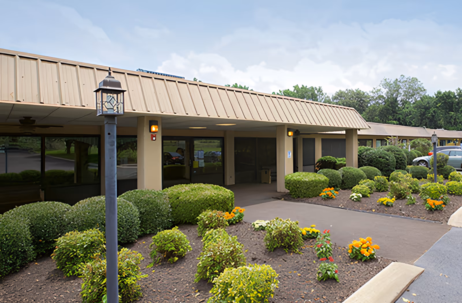 Exterior view of Ahc West Tennessee Transitional Care facility showing the entrance with a covered walkway, surrounded by neatly trimmed bushes and flower beds with orange and yellow flowers. A lamp post is visible in the foreground and trees are in the background under a partly cloudy sky.