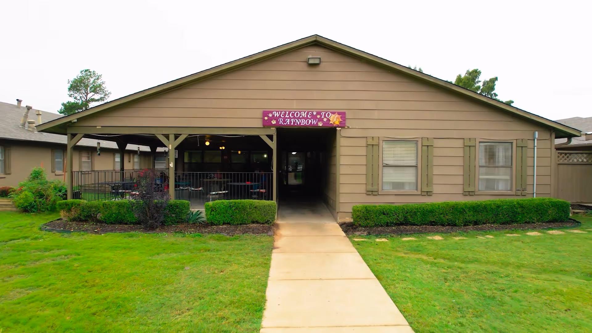 Exterior view of a single-story building with a covered patio area and a walkway leading to an entrance. The building has beige siding, green shutters on the windows, and a sign above the entrance that reads 'WELCOME TO RAINBOW'. There is a well-maintained lawn and bushes in front of the building.