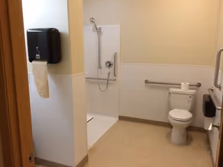 A clean and accessible bathroom with a toilet, a grab bar on the wall, a toilet paper roll on the tank, a wall-mounted soap or paper towel dispenser, and a walk-in shower with a handheld showerhead and grab bars.