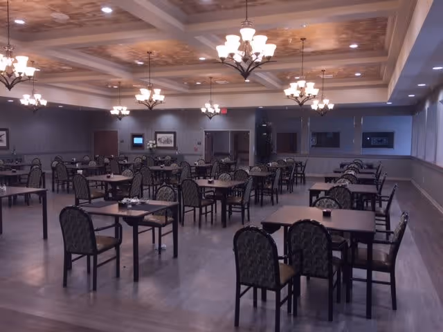 A spacious dining room with multiple square tables and patterned chairs arranged neatly. The ceiling features decorative panels and multiple chandeliers providing warm lighting. The room has a wooden floor and neutral-colored walls with framed artwork.
