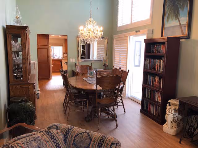 A dining room with a wooden table and chairs under a chandelier, bookshelves, sliding glass door, and an open doorway to the kitchen.