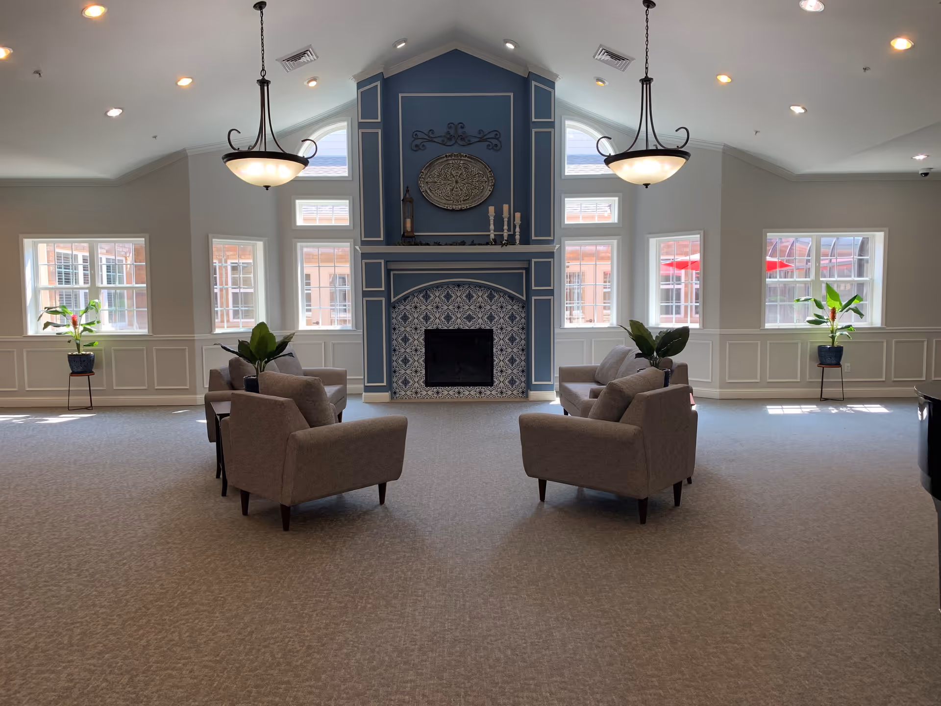 A spacious, well-lit common area with a high vaulted ceiling and two hanging light fixtures. The room features a central fireplace with decorative blue and white tile and a blue accent wall. Four gray armchairs are arranged in two pairs facing each other around the fireplace. There are several windows letting in natural light, and potted plants are placed near the windows.