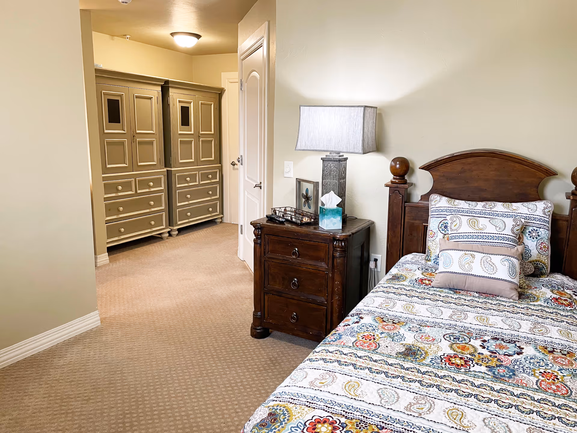 A cozy bedroom with a single wooden bed featuring a colorful patterned bedspread and matching pillows. Next to the bed is a dark wooden nightstand with a lamp, tissue box, and a small decorative item. In the background, there is a hallway leading to a set of beige wardrobes with multiple drawers and cabinet doors. The room has beige carpet and light-colored walls.