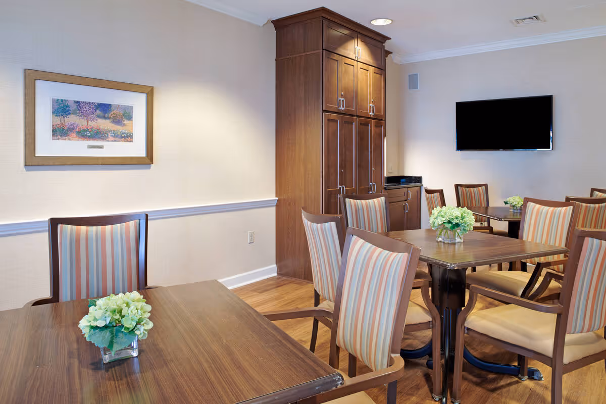 A bright dining room with wooden tables and striped cushioned chairs, small floral centerpieces, a wall-mounted TV, and tall wooden cabinetry.