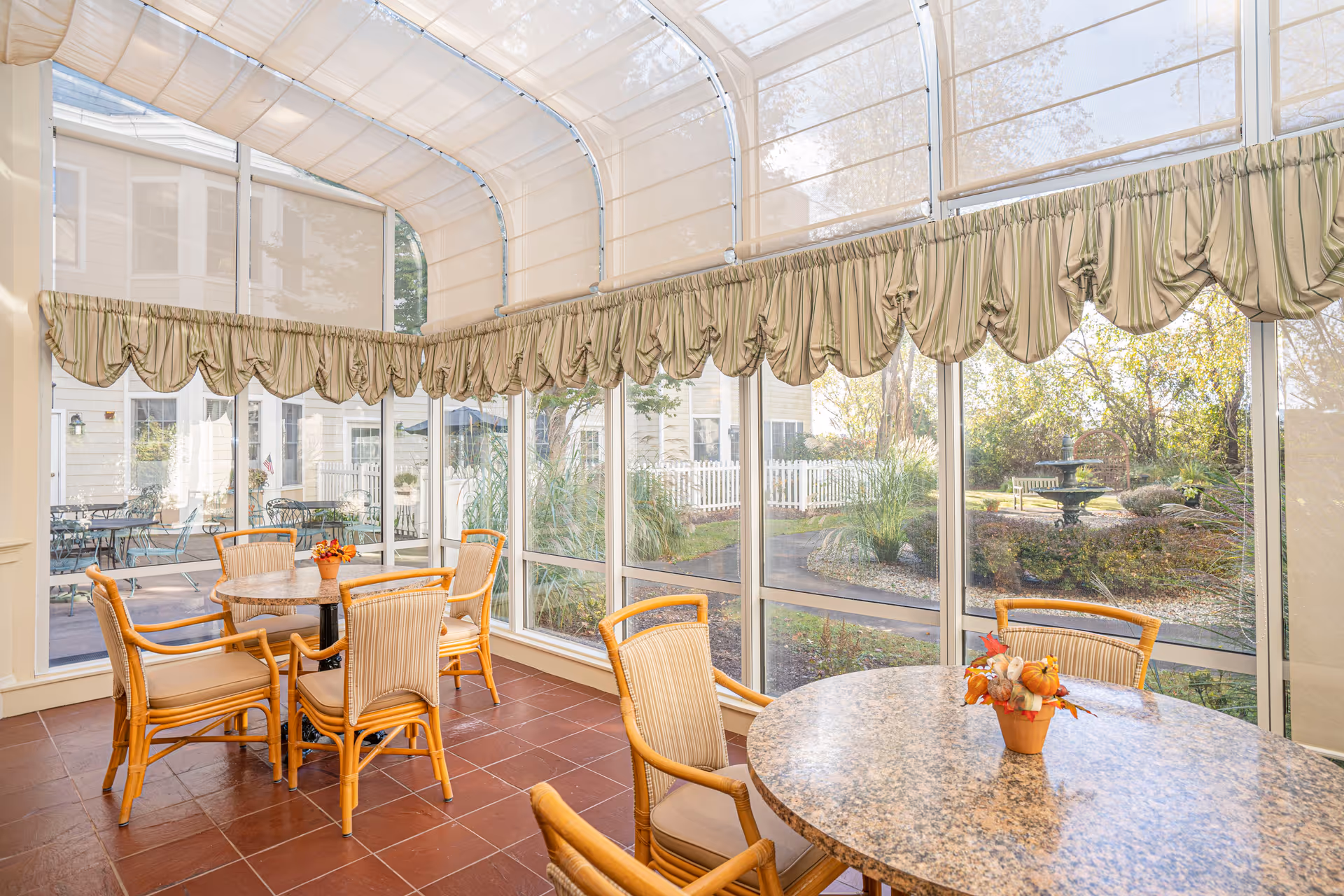 A bright sunroom with large windows and a curved glass ceiling, furnished with round tables and wicker chairs. Outside the windows, a garden with greenery, a white picket fence, and a water fountain is visible. The room has tiled flooring and decorative valances on the windows.