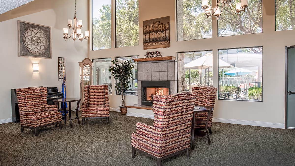 A cozy living room area with four patterned armchairs arranged around two small round tables. There is a fireplace with a fire burning, large windows letting in natural light, a grandfather clock, a piano, and decorative wall art. Outside the windows, patio umbrellas and greenery are visible.