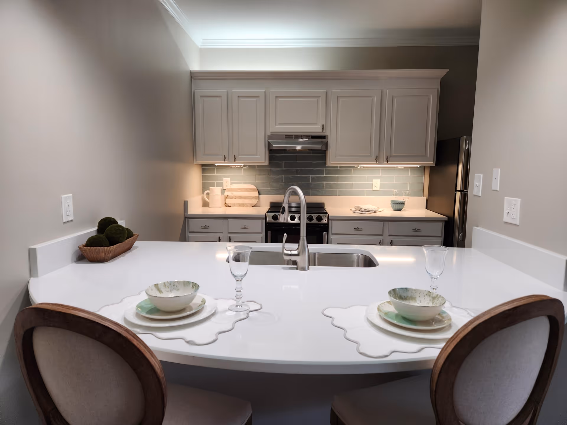 Bright modern kitchen with a white island countertop and sink, two place settings and chairs in the foreground and cabinets and stove in the background.